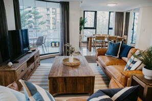 A living room with a leather sofa, a wooden coffee table, a television on a wooden stand, and a dining area with a table and chairs. - Biarritz by the Sea | Resort Days & Ocean Views (Gold Coast)