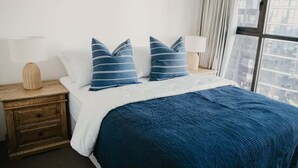 En säng med ett blått täcke, två kuddar, ett träbord vid sängen med en lampa och en utsikt över byggnader genom ett fönster.