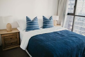 A bed with a blue quilt, two pillows, a wooden nightstand with a lamp, and a view of buildings through a window. - Biarritz by the Sea | Resort Days & Ocean Views (Gold Coast)