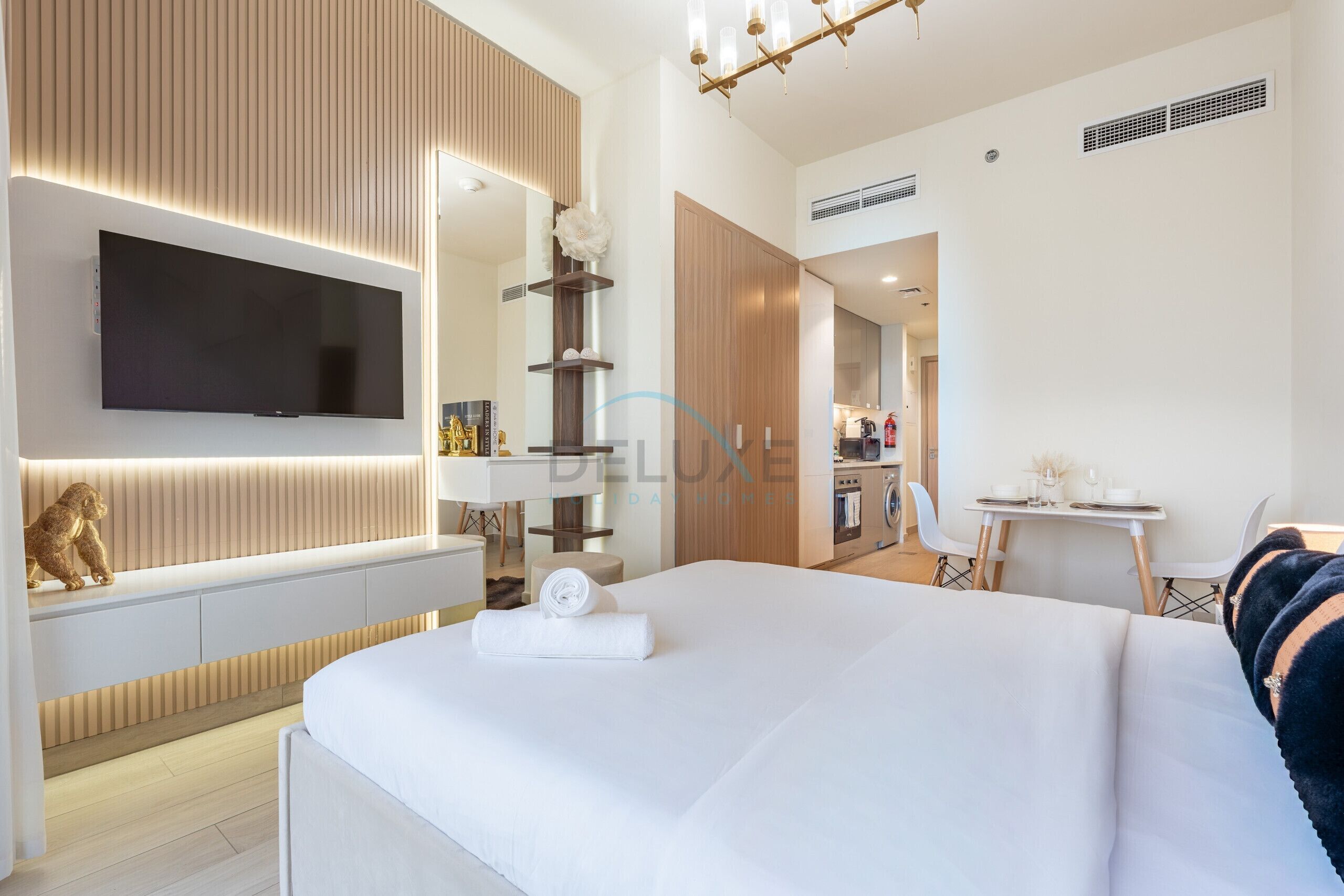 Una habitación de hotel moderna con una televisión de pantalla plana, una cama con una toalla enrollada, un escritorio con una silla y una kitchenette.