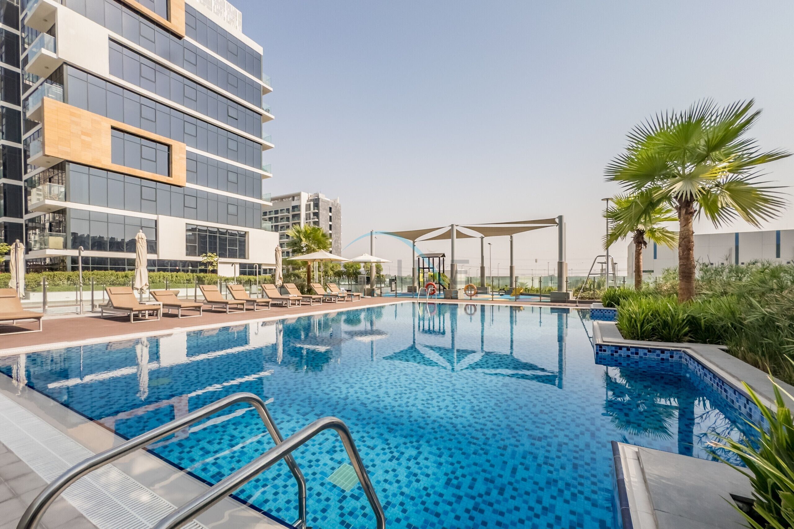 Ein Pool mit Liegestühlen und Sonnenschirmen, umgeben von modernen Gebäuden und Palmen.