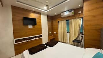 A hotel room with a bed, a flat-screen TV mounted on the wall, an air conditioning unit, a ceiling fan, and a window with curtains.