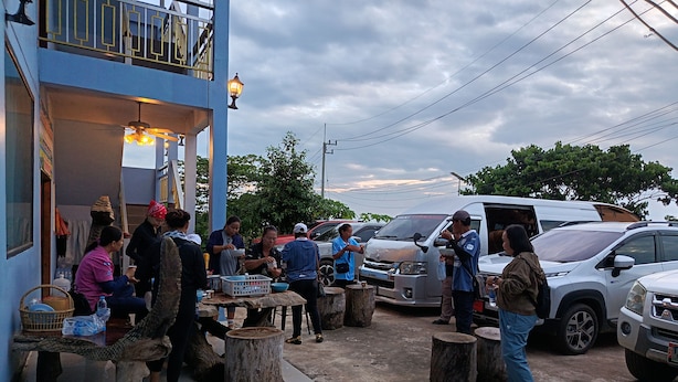 A group of people gathered around a table outside a building with a balcony, with parked cars and a cloudy sky in the background.