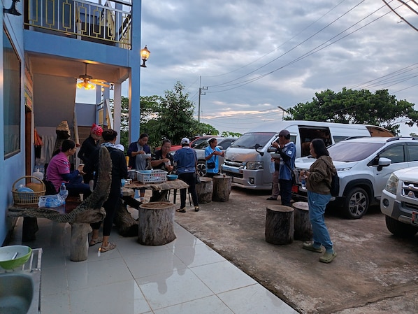 A group of people gathered around a table outside a building with a balcony, with parked cars and a cloudy sky in the background.