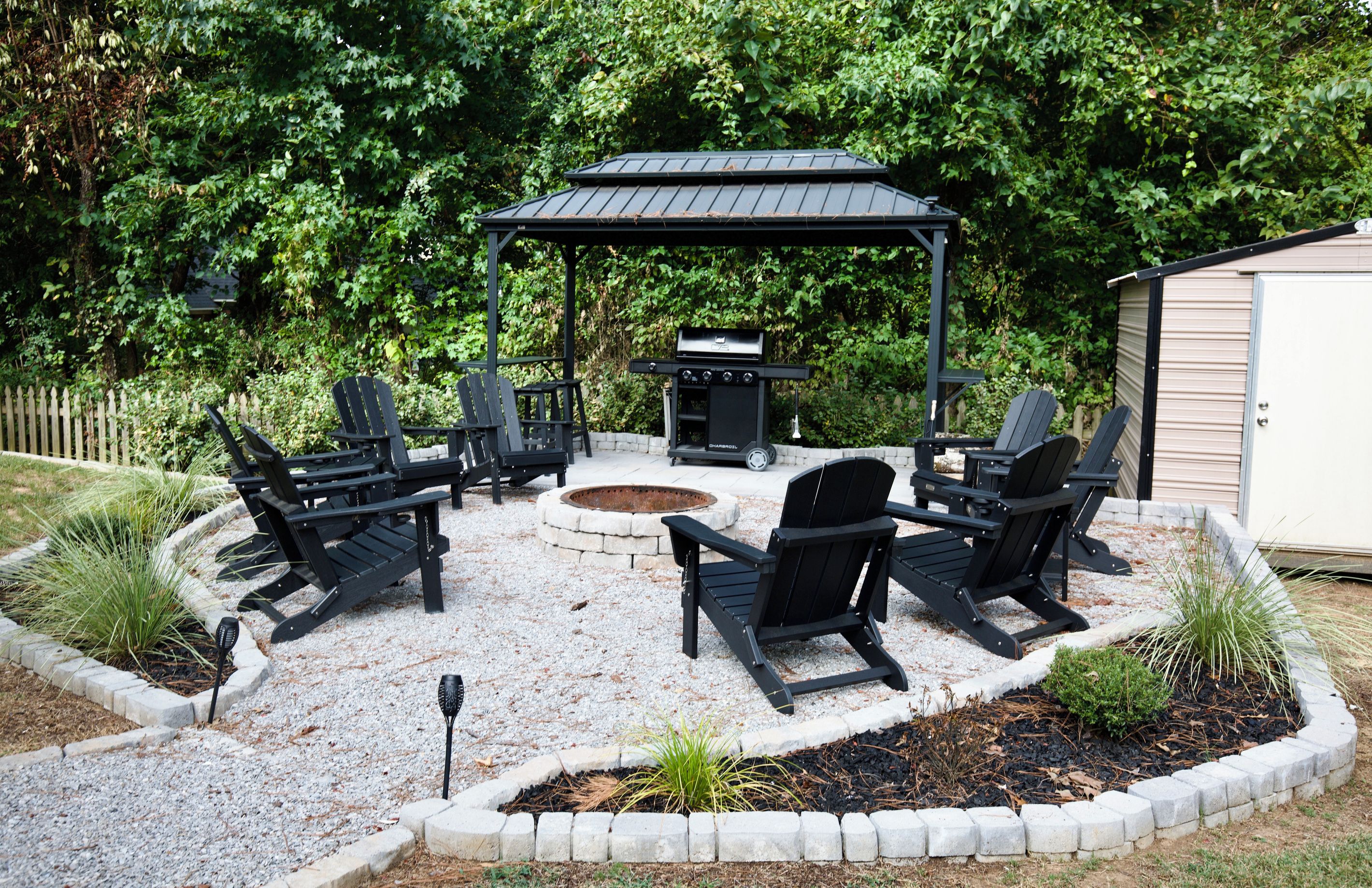 A circular outdoor seating area with Adirondack chairs and a fire pit, surrounded by a gravel patio and a stone border.