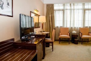 Une chambre d’hôtel équipée d’une télévision, d’un bureau, de deux chaises, d’une lampe et d’une grande fenêtre avec des rideaux.