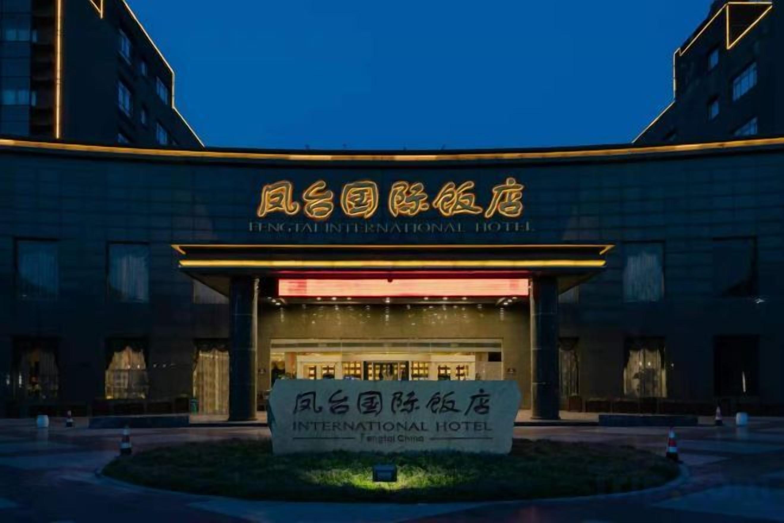 A hotel entrance with illuminated signage in a foreign language, a well-lit facade, and a landscaped area in front.