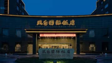 A hotel entrance with illuminated signage in a foreign language, a well-lit facade, and a landscaped area in front.