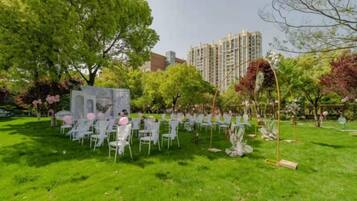 An outdoor seating area with white chairs arranged in rows, a stone structure, and trees in the background.