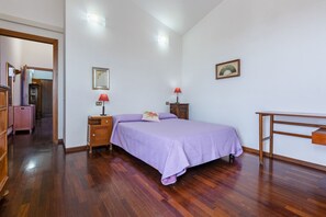 Ein Schlafzimmer mit Holzboden, einem Bett mit violetter Bettdecke, einem Nachttisch mit Lampe, einer Kommode und einem Schreibtisch mit Stuhl.
