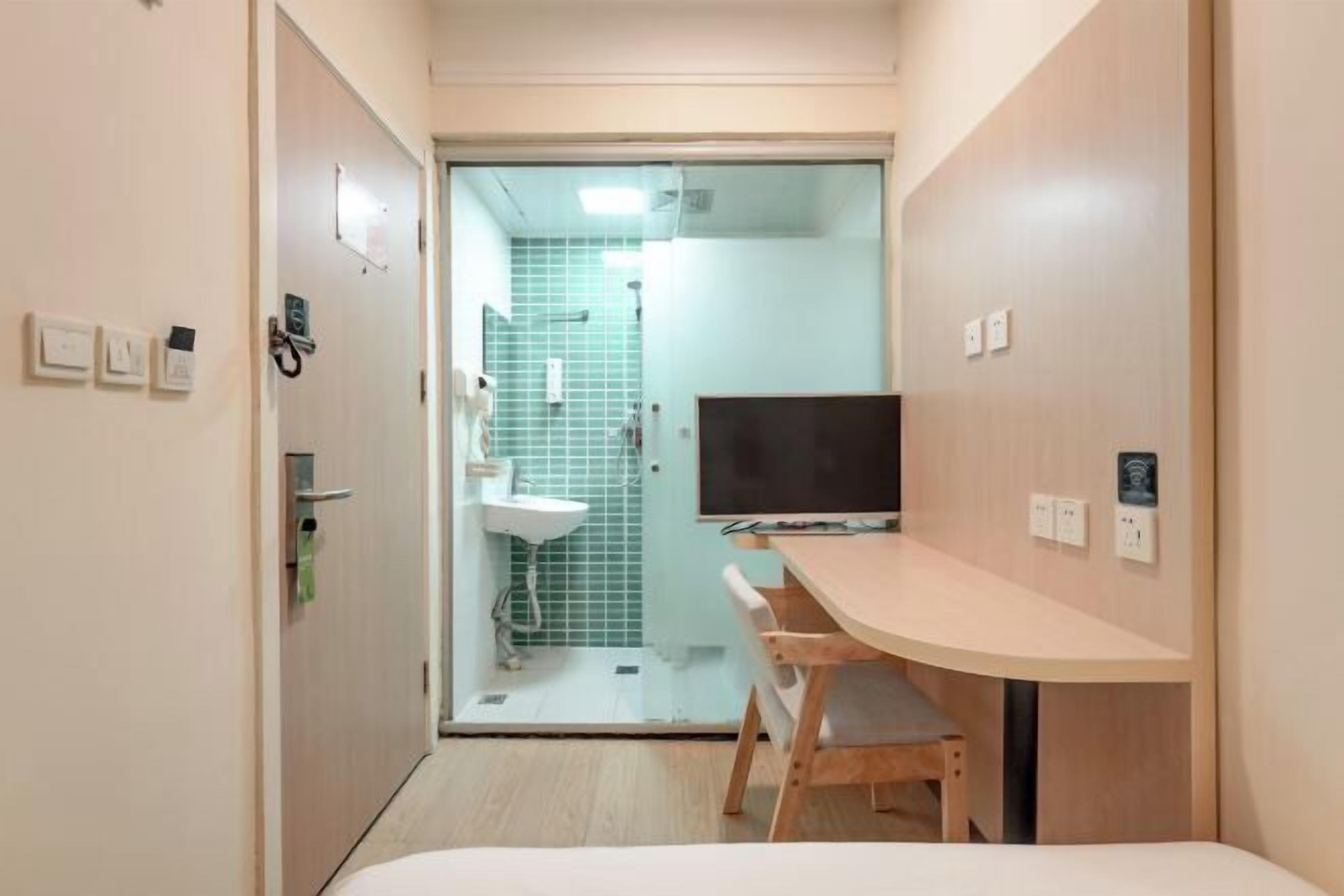 A compact hotel room with a bed, a desk, a chair, a television, and a bathroom with a shower and sink.