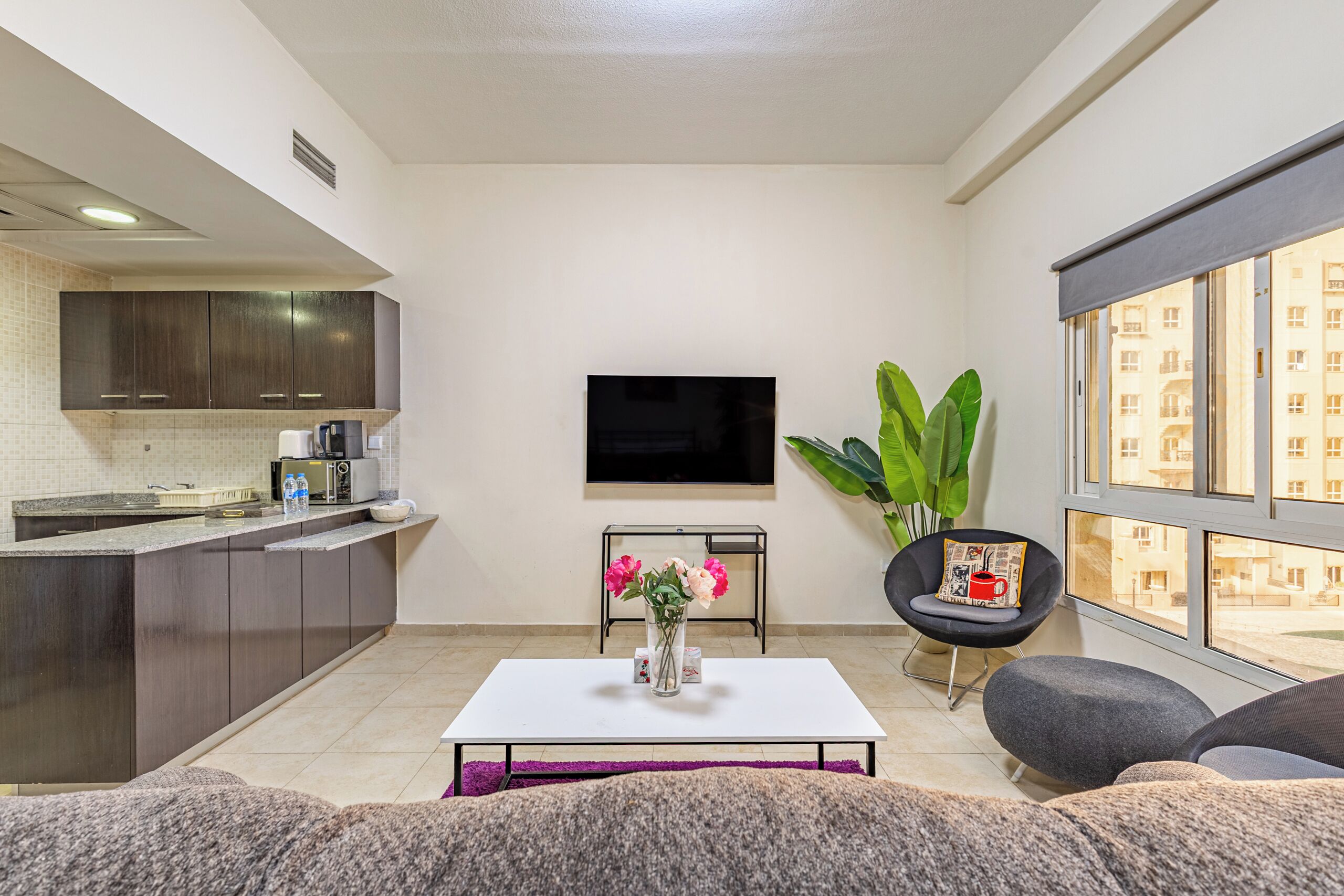 A modern living room with a kitchen area, a flat-screen TV mounted on the wall, a coffee table with flowers, and a large window with a view of buildings.