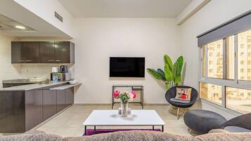 A modern living room with a kitchen area, a flat-screen TV mounted on the wall, a coffee table with flowers, and a large window with a view of buildings.