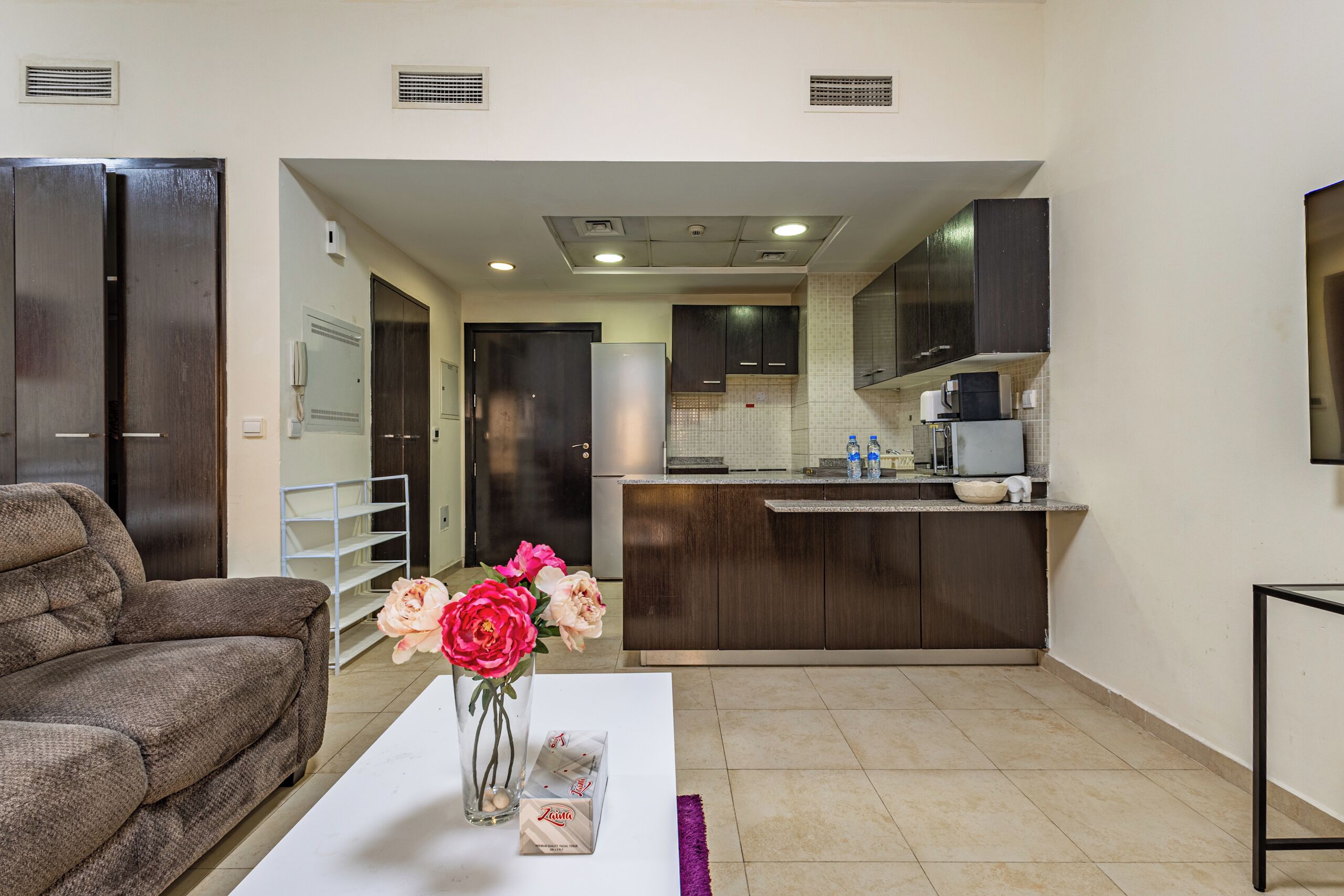 A living room with a sofa, a coffee table with a vase of flowers, and a kitchen area with cabinets and a refrigerator.