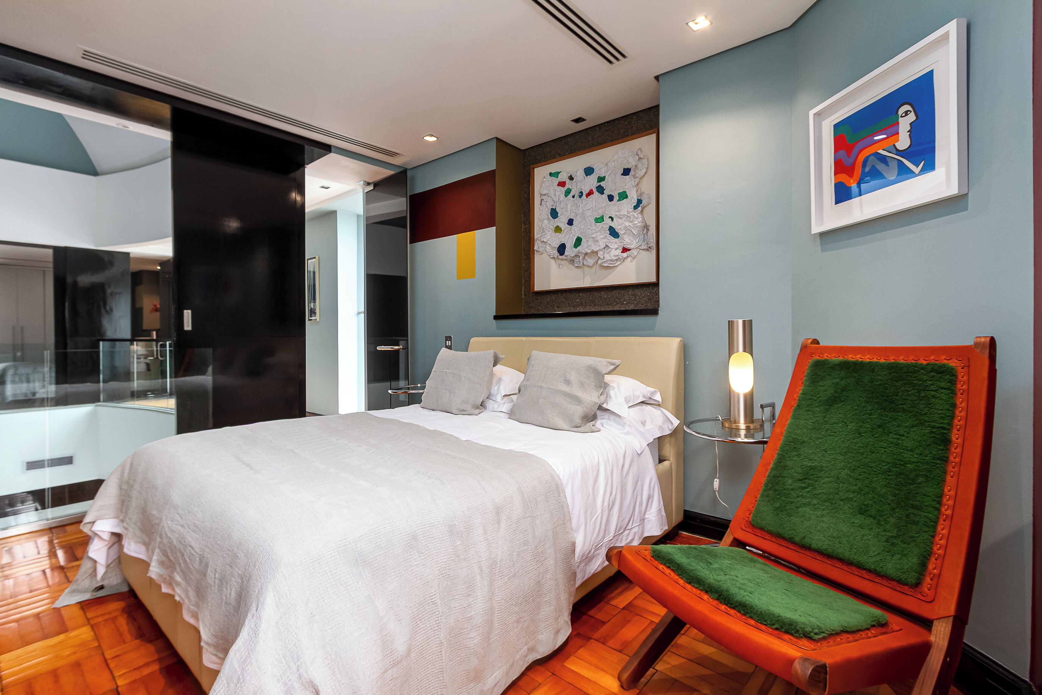 A modern bedroom with a large bed, a bedside table with a lamp, a chair with a green cushion, and artwork on the walls.