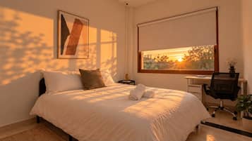 A bedroom with a bed, a desk, a chair, and a window with a view of trees and a sunset.