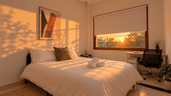 Un dormitorio con cama, escritorio, silla y ventana con vista a árboles y al atardecer.