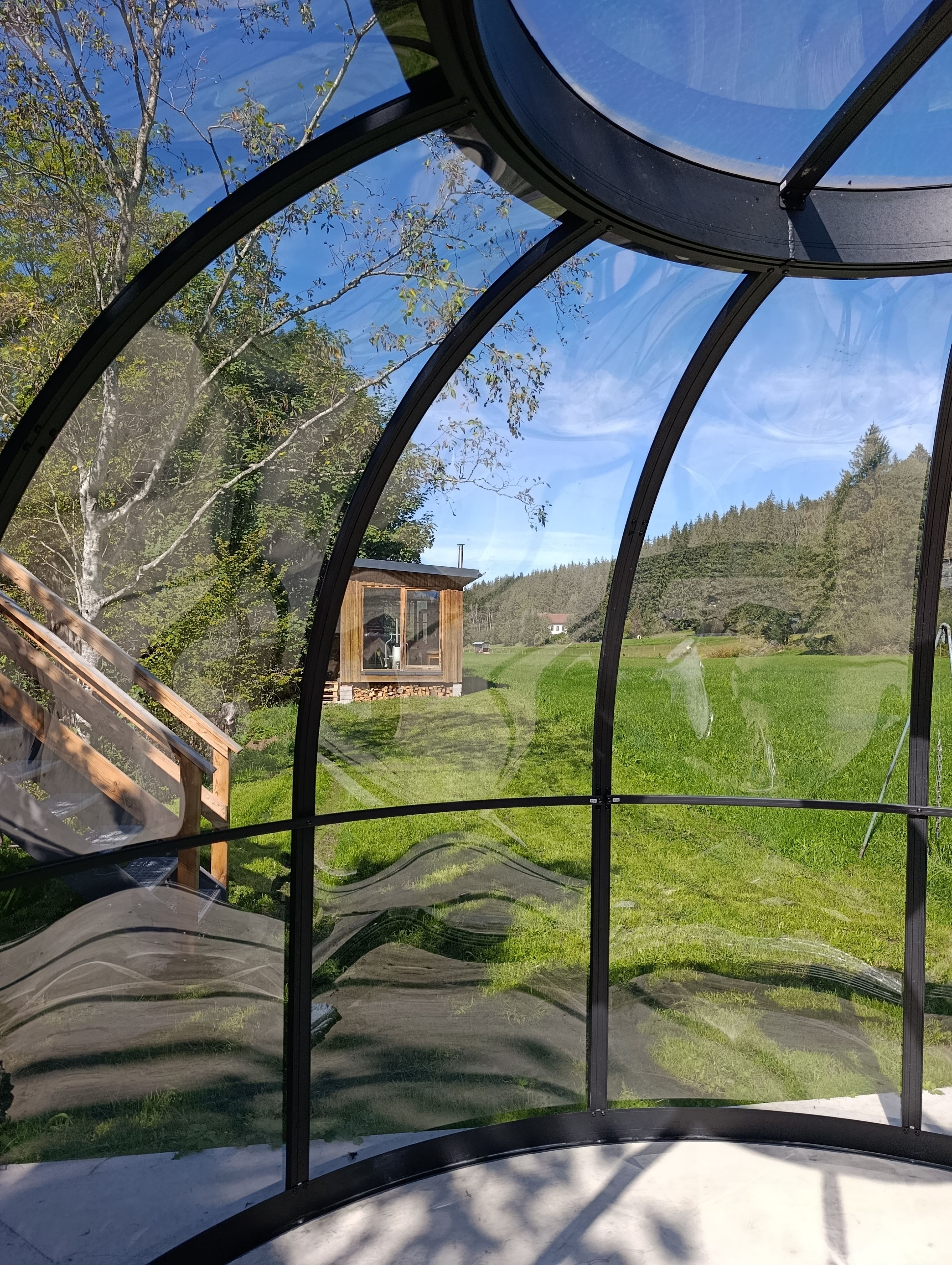 Blick durch eine gebogene Glaskonstruktion auf eine Holzhütte und eine grüne Landschaft.