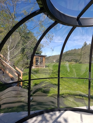 A view through a curved glass structure with a wooden cabin and a grassy landscape.