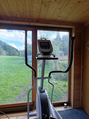 An exercise bike with a digital display, positioned in a room with wooden walls and a large window overlooking a green landscape.