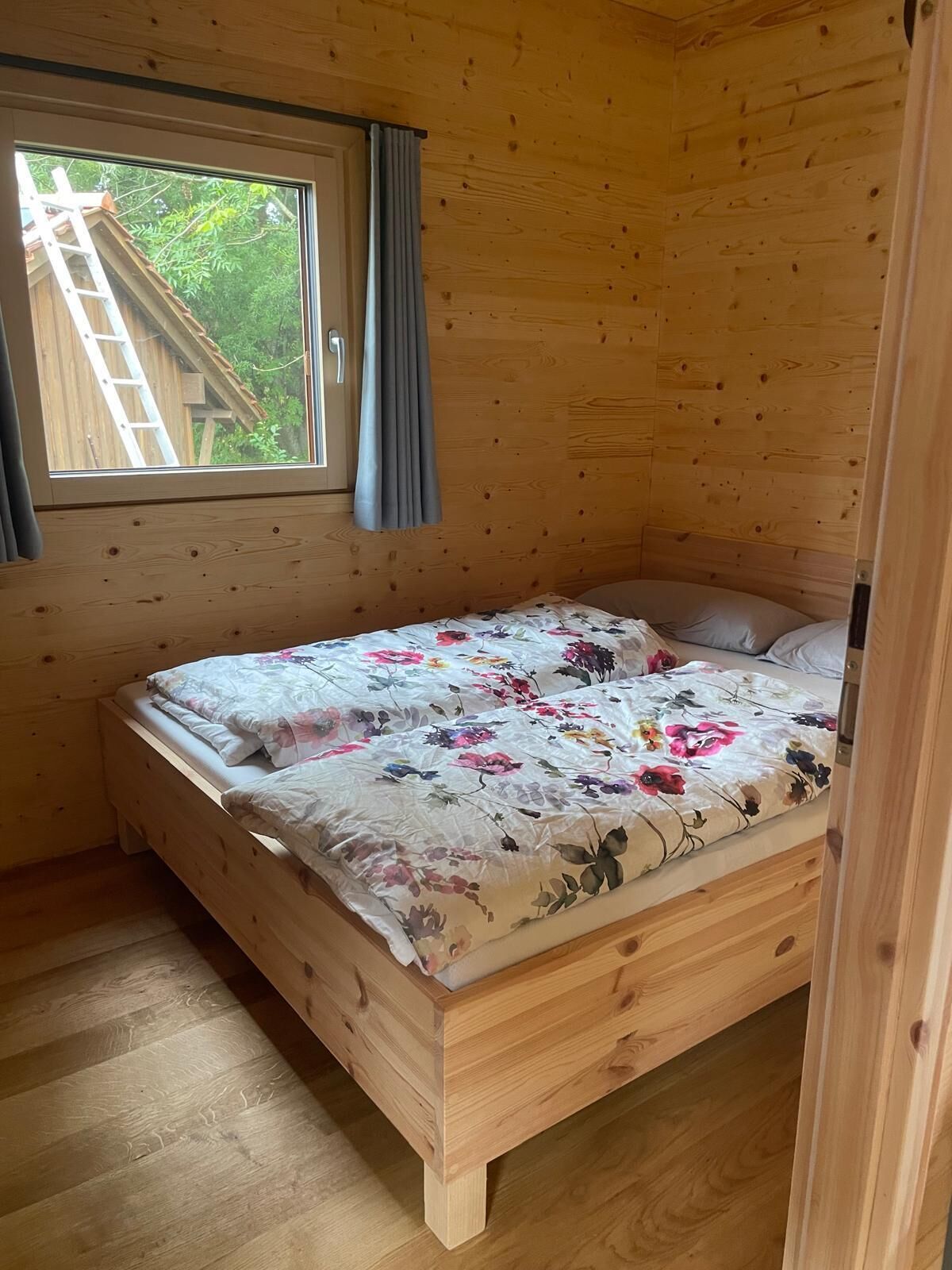 A wooden room with a bed, a window, and a view of greenery outside.