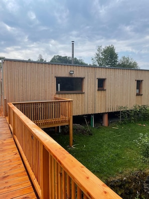 A wooden deck leading to a modern building with a metal pole, surrounded by grass and trees.