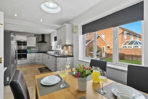 A modern kitchen with white cabinets, a dining area with a wooden table, and a view of a brick house through large windows. - Spacious 5 Bed 2 Bath House • Driveway Parking • Near NEC • Solihull • Airport (Balsall Common)