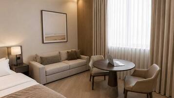 A hotel room with a bed, a sofa, a desk with a laptop, and a dining table with chairs.