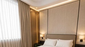 Una habitación de hotel moderna con una cama grande, una mesita de noche y una banca, además de un ventanal con cortinas.