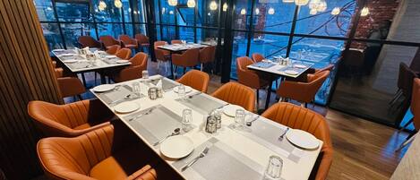 A restaurant with tables set for dining, large windows, and hanging light fixtures.
