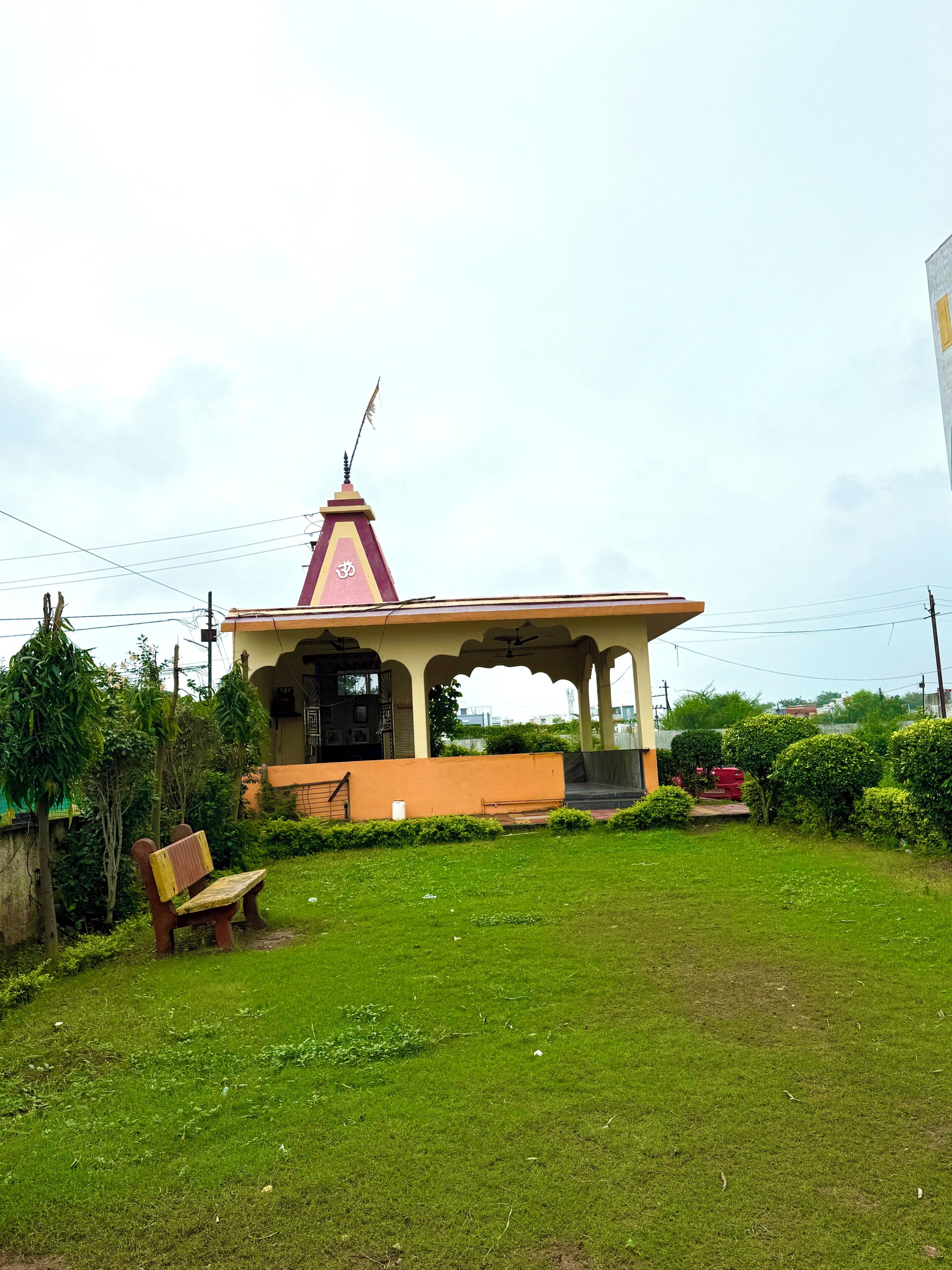 A small, single-story building with a flat roof and a central tower, surrounded by a grassy area with a bench and a red vehicle in the background.