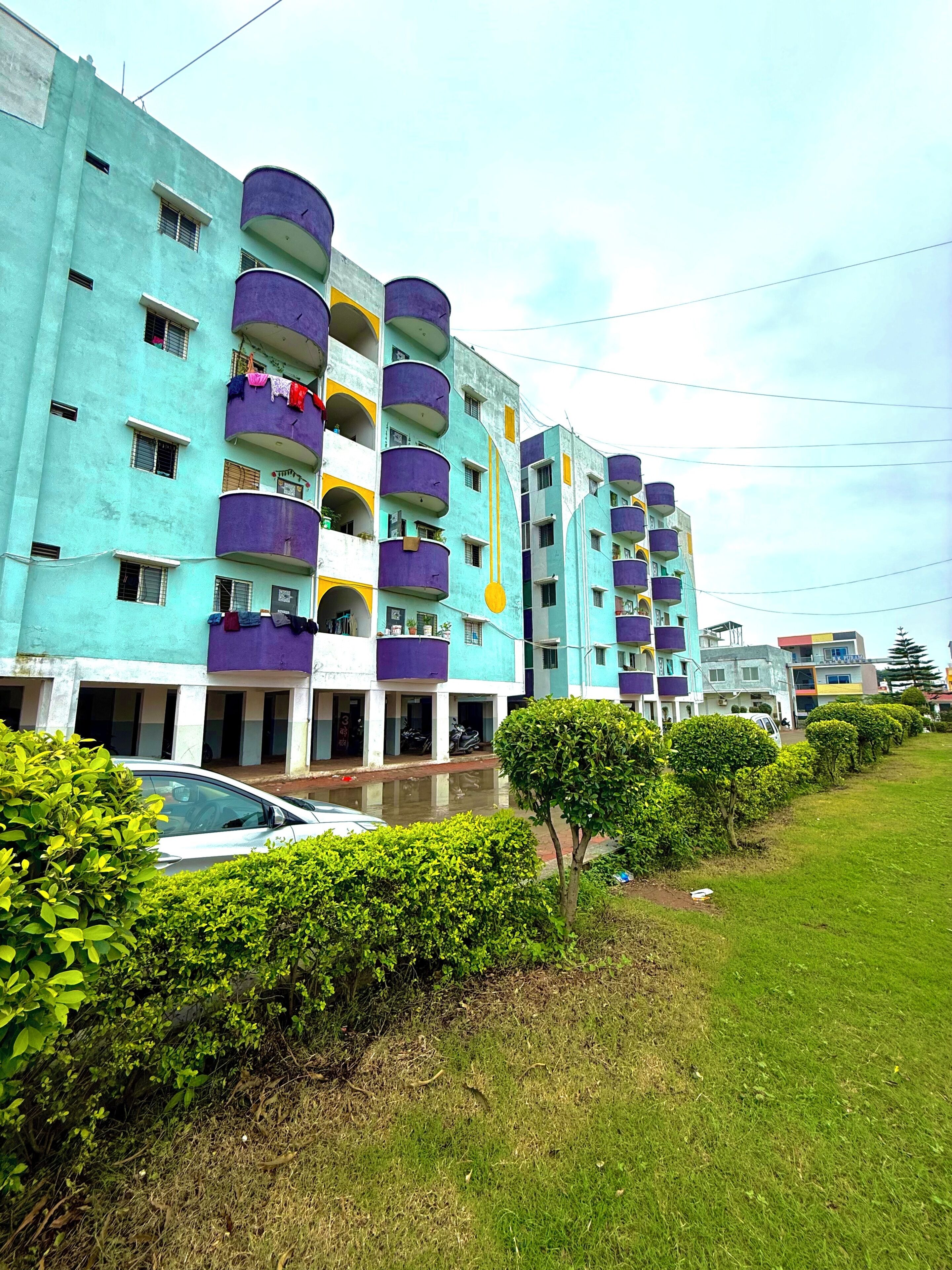 A multi-story residential building with balconies, a car parked in front, and a well-maintained lawn.