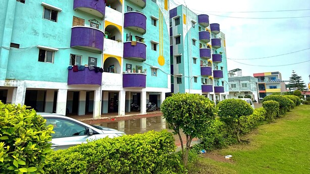 A multi-story residential building with balconies, a car parked in front, and a well-maintained lawn.