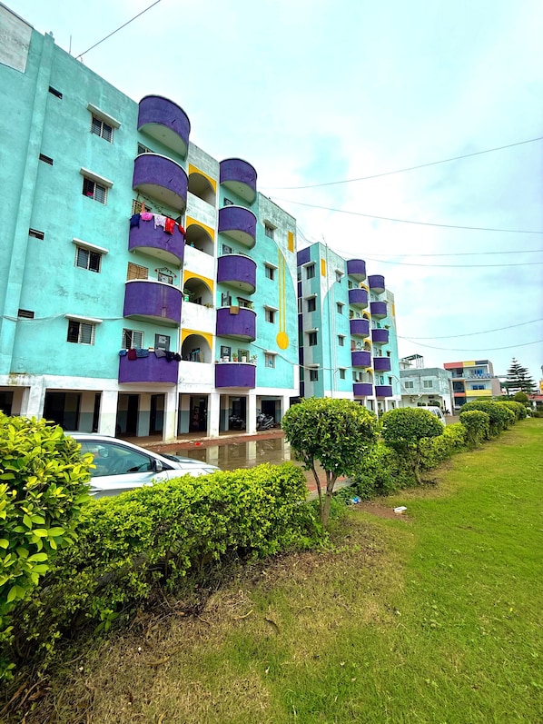 A multi-story residential building with balconies, a car parked in front, and a well-maintained lawn.
