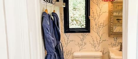 Une petite salle de bain avec des toilettes, une fenêtre, un lavabo et deux peignoirs suspendus à des crochets.