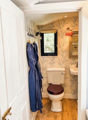A small bathroom with a toilet, a window, a sink, and two robes hanging on hooks.