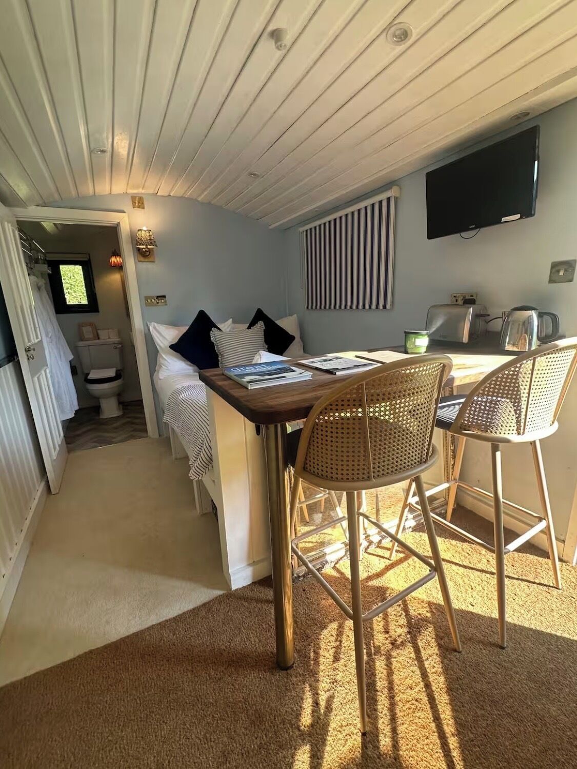 A compact room with a bed, a dining table with chairs, a television mounted on the wall, and a window with blinds.