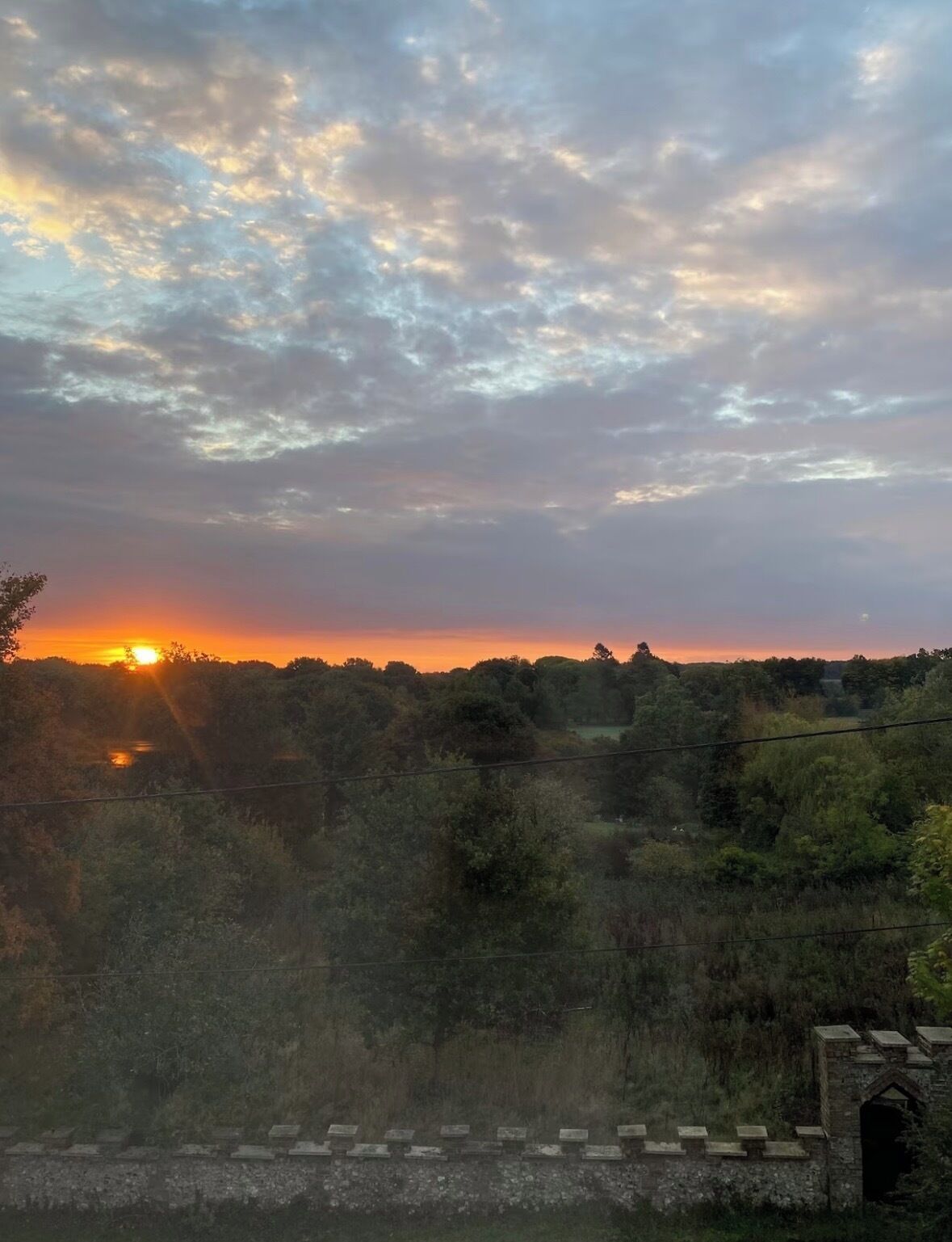 Een zonsondergang boven een bebost landschap, met een stenen brug op de voorgrond.