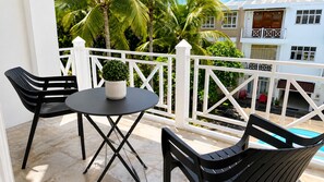 A balcony with a round table and two chairs, a potted plant, and a view of a pool and buildings.