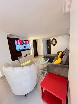 Un saló modern amb televisió plana, un sofà gris amb coixins grocs, una butaca blanca i una taula auxiliar vermella.