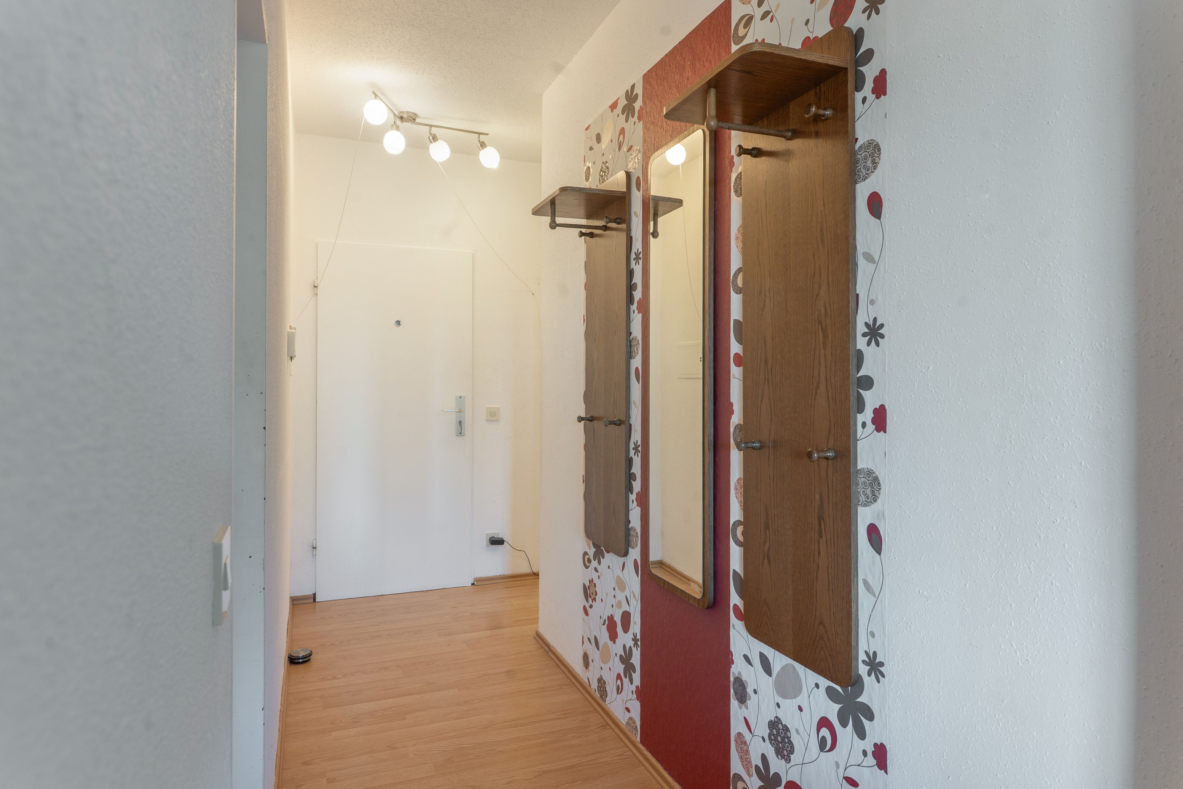 A hallway with wooden flooring, a white door, and a mirror with floral wallpaper.