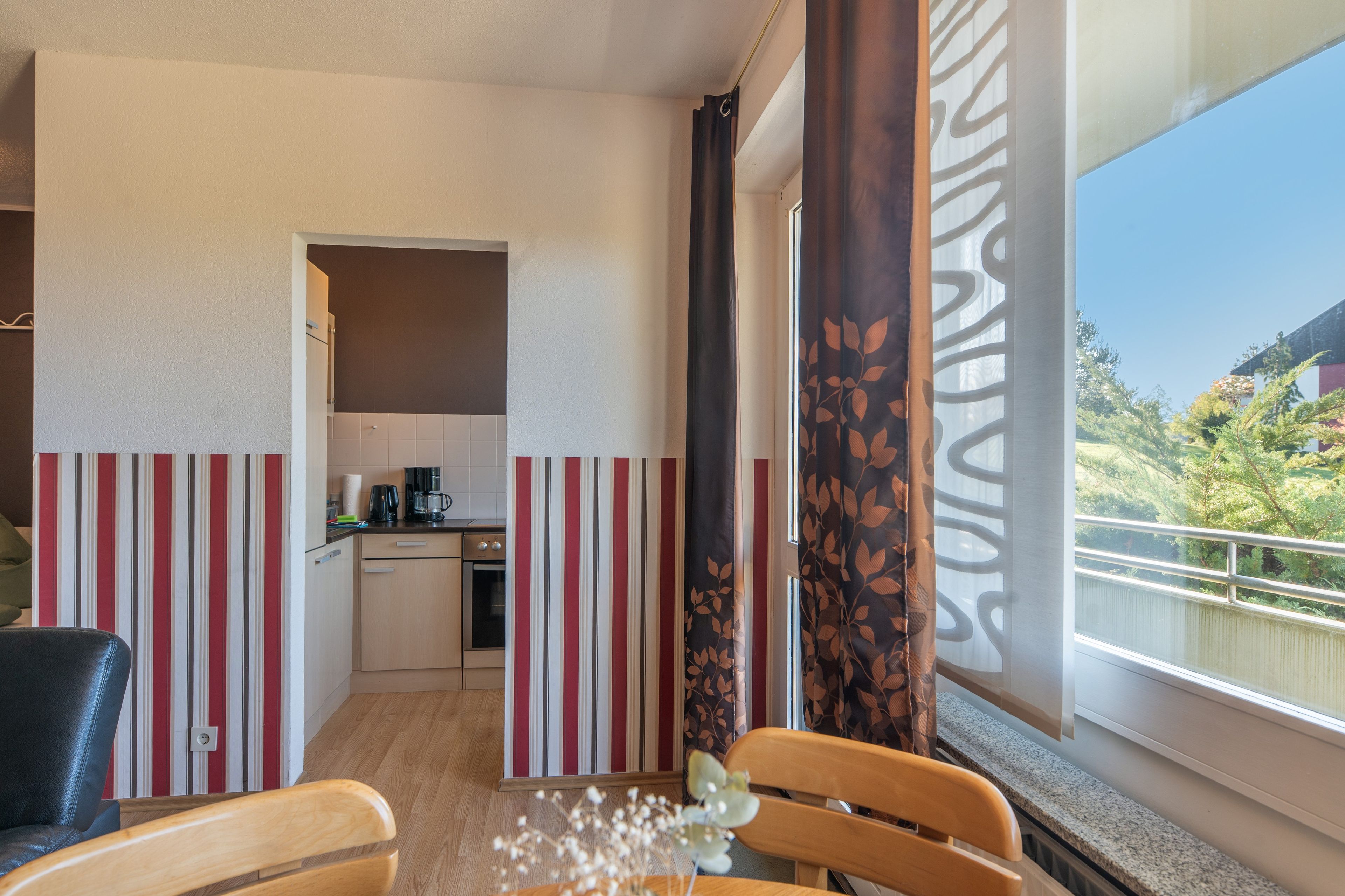 Une cuisine compacte équipée d’une machine à café, un coin repas avec un papier peint à rayures et une vue sur la verdure à l’extérieur.