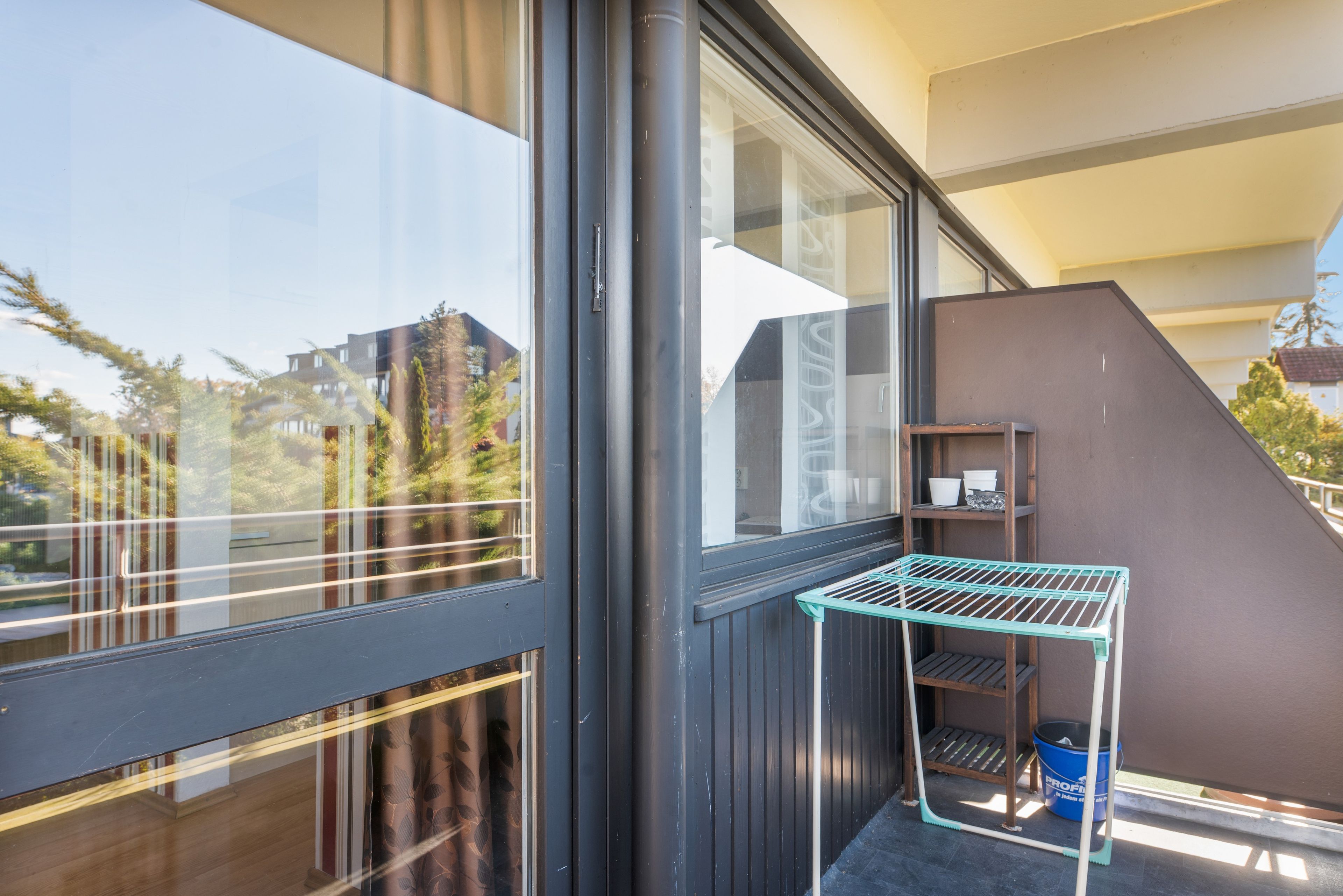 Ein Balkon mit einem Metallregal und einem blauen Eimer, mit Blick auf ein Wohngebiet.