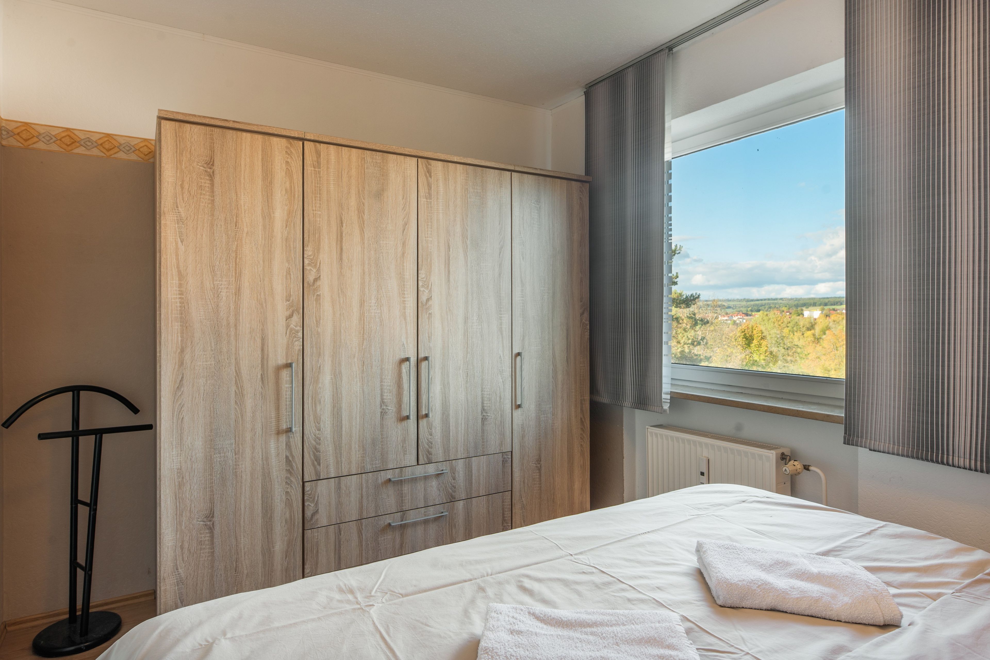 Ein Schlafzimmer mit großem Kleiderschrank, einem Bett mit weißer Daunendecke und Blick auf eine Landschaft durch ein Fenster.