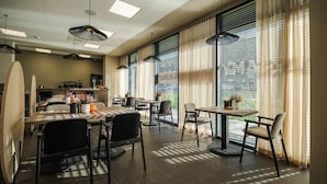 A modern dining area with multiple tables and chairs, large windows with blinds, and pendant lighting.