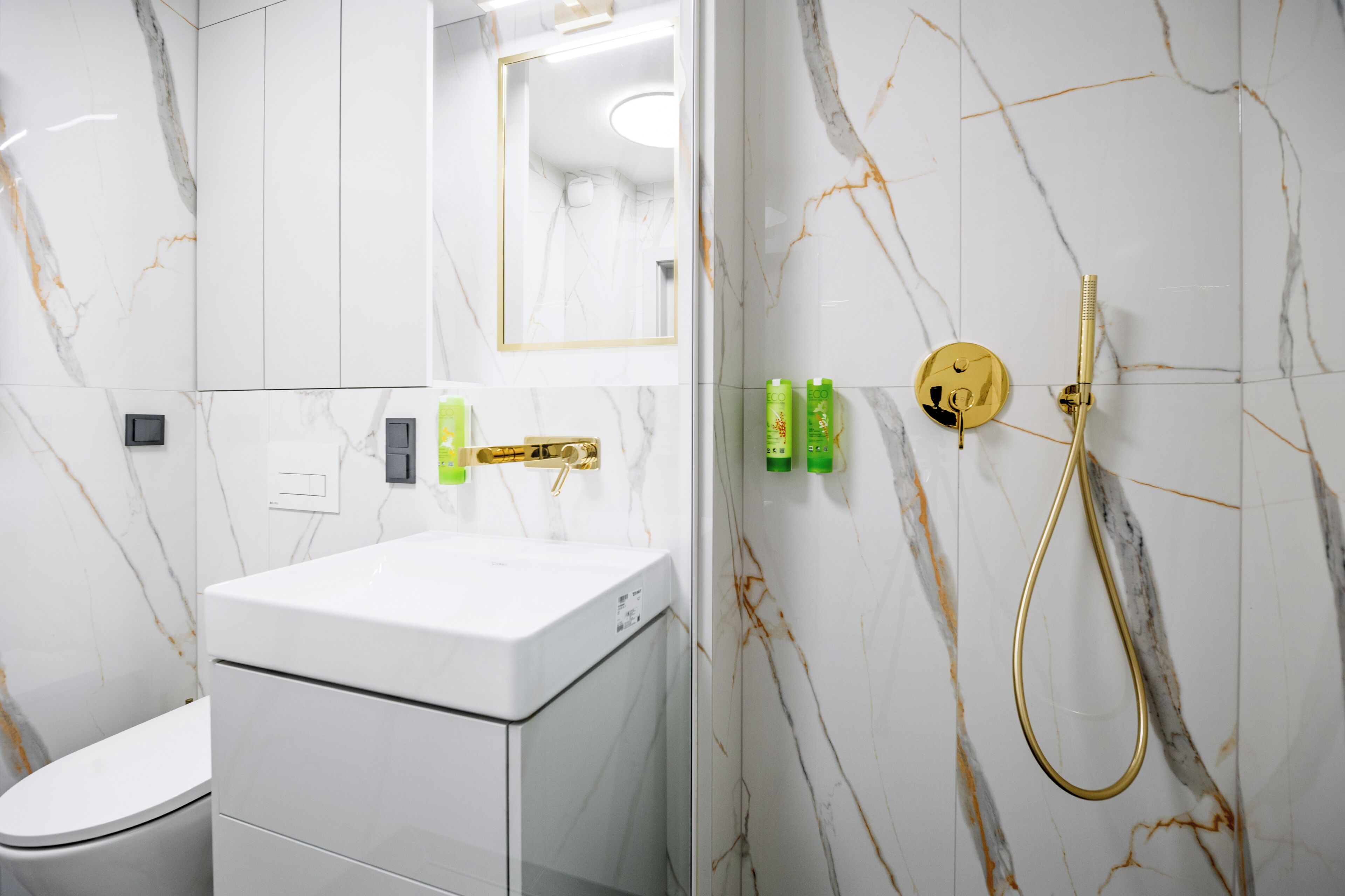 Un baño moderno con un lavabo blanco, un inodoro y una ducha con cabezal dorado.
