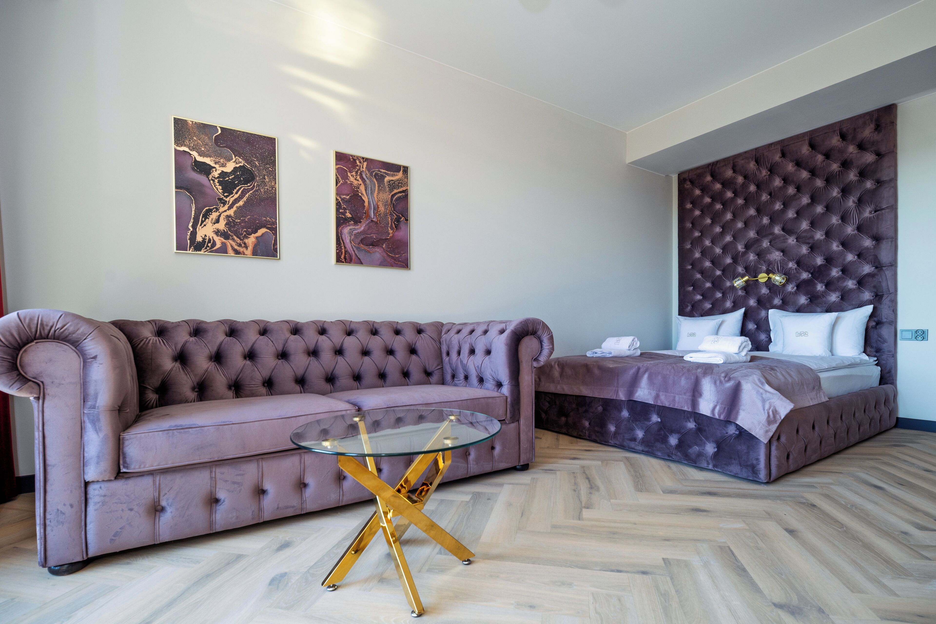 A modern hotel room with a tufted sofa, a glass coffee table, a large bed with a tufted headboard, and two abstract paintings on the wall.