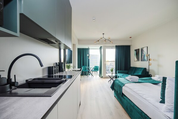 A modern hotel room with a kitchenette, a bed with green bedding, a dining area with a table and chairs, and a balcony with curtains. - K52 Apartments (Wrocław)