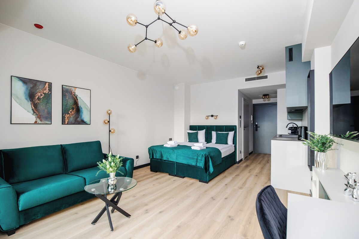 Una habitación de hotel moderna con un sofá verde, una cama con ropa de cama verde, una mesa de centro de vidrio y una lámpara de techo original.