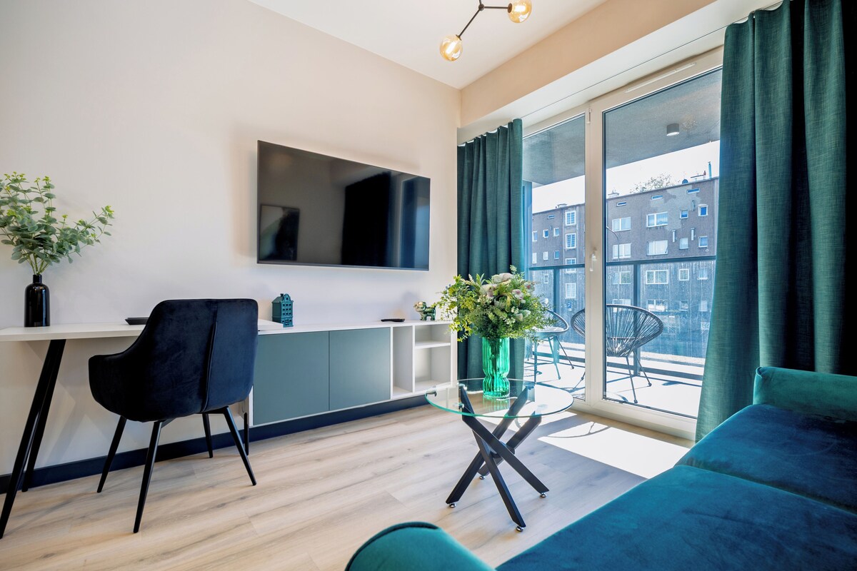 Una sala moderna con un sofá, un televisor montado en la pared, una mesa de comedor con sillas y un ventanal con cortinas.